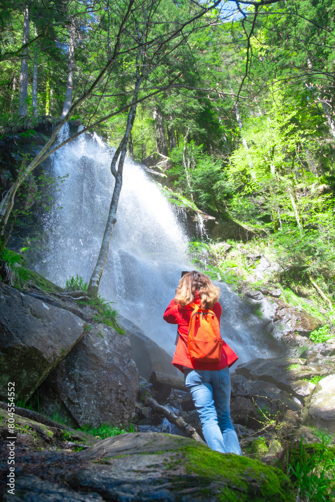 Naklejka premium hiker woman in a red jacket is taking photos under a waterfall in the forest