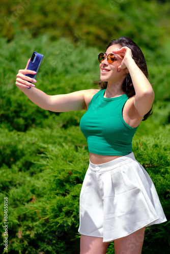 Beautiful girl in a green top and white shorts walks in a city park on a sunny summer day. Taking a selfie, laughing while holding a mobile phone