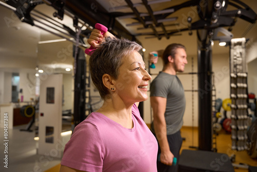 Middle aged woman and blur trainer doing exercise with dumbbells for shoulders