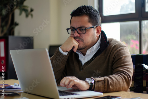 Seated at his office desk, a busy Latin business man is engrossed in his laptop. The professional businessman, a dedicated employee of the company, carefully examines the computer screen,