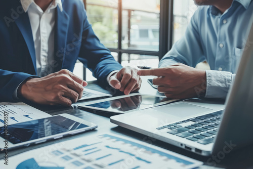 Two business men, constituting a busy executive team, are deep in discussion regarding a digital project and corporate strategy while utilizing a laptop and tablet in the office. One functions as a photo