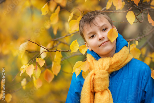 A teenager in a blue jacket and yellow scarf playing hide and seek behind a yellow leaf against the backdrop of autumn nature.
