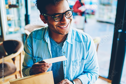 Cropped image of handsome cheerful businessman in stylish eyewear sincerely happy about content of letter indoors.Mature smiling male person enjoying time sitting at wooden table in cozy cafe