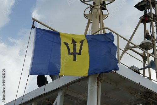 Flying Barbados flag, vertical triband of ultramarine and gold with the black trident-head, on the wind hanging on navigational mast near funnel of merchant container vessel when mooring in Bridgetown