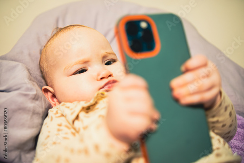Baby is holding a cell phone in its hand, looking at the screen with curiosity, showing the contrast between the innocence of childhood and the modern technology.