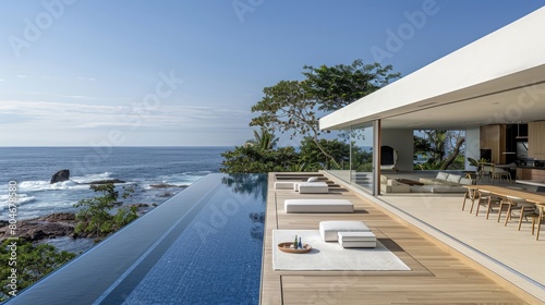 A large house with a pool and a balcony overlooking the ocean. The pool is surrounded by white lounge chairs and a dining table is set up on the balcony. The house has a modern