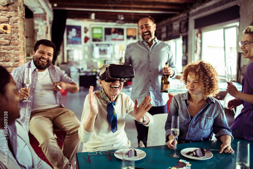 Diverse group of people laughing with senior woman using VR headset at office party photo