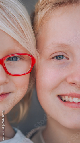 A close-up portrait of two blond sisters with blue eyes smiling one of them wearing red glasses