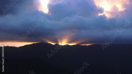 The sun shines brightly through the clouds, casting warm glow over the majestic mountains in Kualoa, Oahu, Hawaii, USA.The clouds move slowly across the sky, creating dynamic play of light and shadow.
