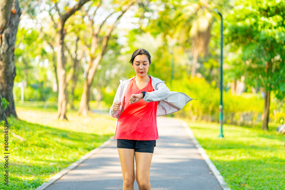 Asian woman using smart watch checking heart rate and calories burned during jogging exercise at park. Healthy woman do sport training running workout in the city. Health care technology concept. Asian woman using smart watch checking heart rate and calories burned during jogging exercise at park. Healthy woman do sport training running workout in the city. Health care technology concept.