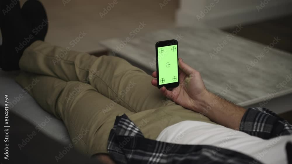 Man resting on a couch at home, using smartphone with green mock-up screen Man resting on a couch at home, using smartphone with green mock-up screen
