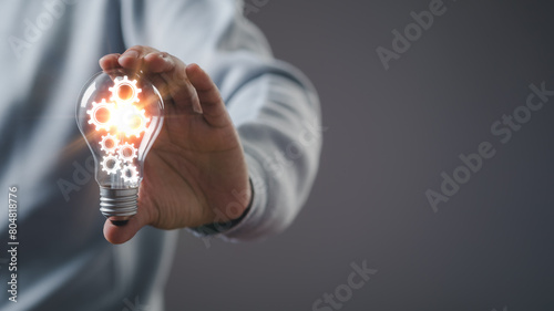 businessman Hand holding smart phone and light bulb with gear glowing with orange light and ray. Creative and new business knowledge thinking idea concept.	
