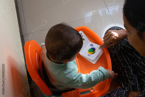 A mother teaching alphabets to her child while making him her eat banana, food. Book, study, learning, focus, chair, feeding tray, table, habit building, development