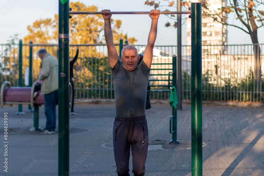 Front view of mature man wearing sports clothes doing pull-ups during a workout on a promenade. Healthy, sporty lifestyles and slimming concept
