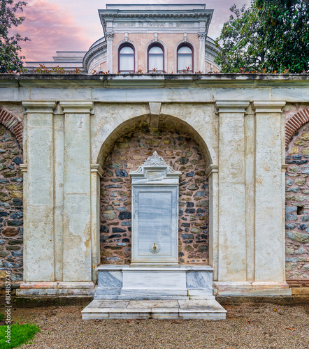 A beautiful white marble drinking fountain in a stone brick wall at the garden of the Mecidiye Pavilion, Istanbul, Turkey, with the palace in the background