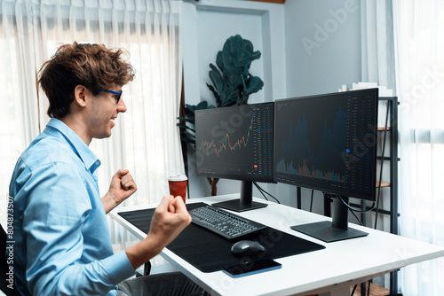 Successful young business trader raising fist up with highest profit stock on global market online data graph on pc screens in real time at modern office, earning valued stock rate investment. Gusher. photo