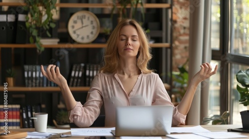 Calm young executive meditating at her desk with laptops and documents around, balancing stress with serenity