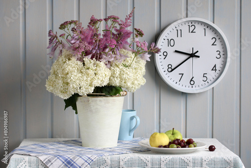 a summer rural still life with a bouquet of hydrangeas and other flowers on the table in the cottage. a white wall clock. summer. apples and gooseberries.