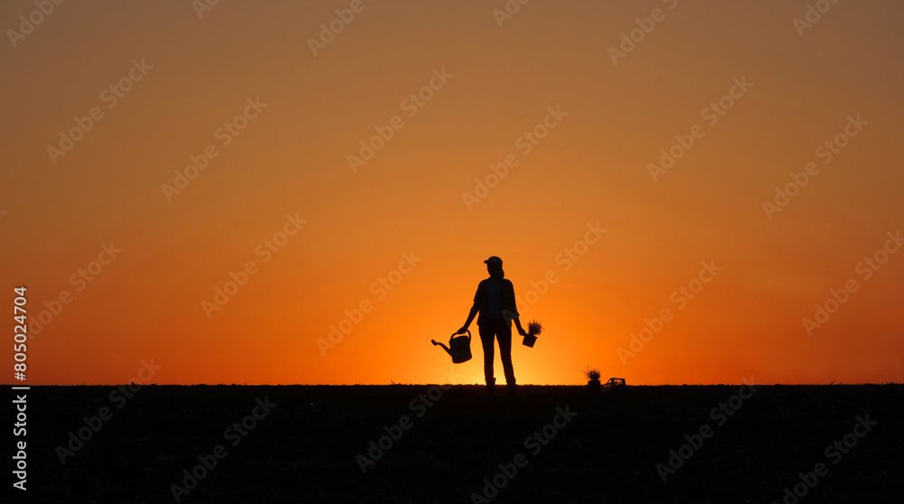 Against the sunset backdrop, the silhouette of a woman farmer holds a waterer and a seedling in her hand, standing in a field.