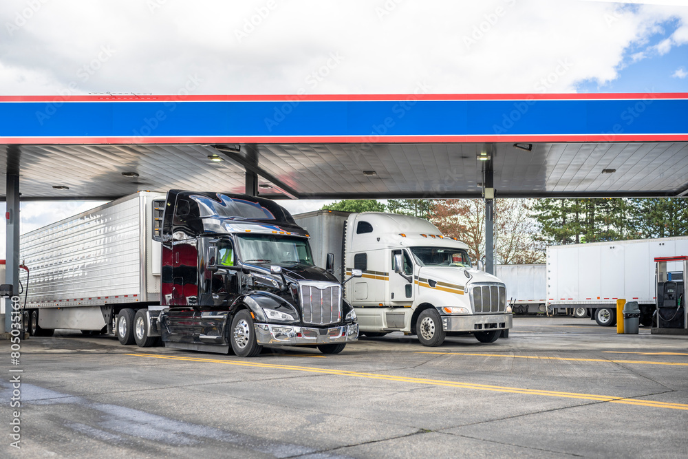 Black and white big rigs semi trucks with semi trailers standing on the truck stop gas station filling tanks with fuel to continue delivering goods to their destination