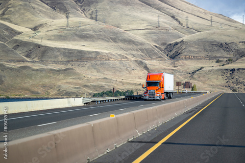 Bright orange bonnet big rig semi-truck with grille guard transporting cargo in dry van semi-trailer climbing uphill on the highway road with bare bald mountains photo