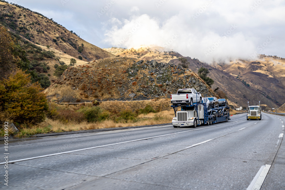 Classic car hauler big rig semi truck transporting cars on the semi trailer climbing uphill on the mountain highway road in California