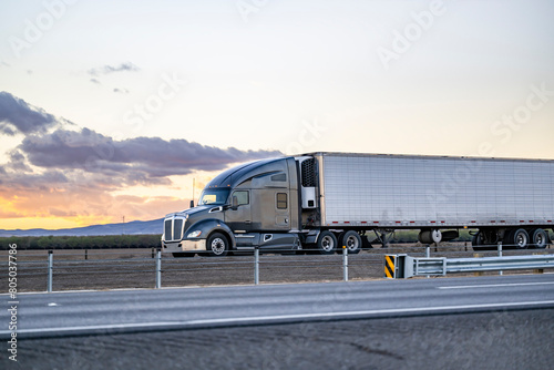 Dark gray big rig semi-truck tractor transporting cargo in refrigerator semi trailer driving on the divided highway road with sunset sky in California © vit