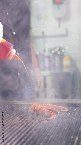 Chef pours sauce on the grilling octopus on an electric grill in the restaurant kitchen in slow motion. Vertical video format.