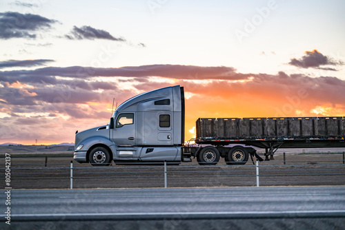 Profile of the gray big rig semi-truck with extended cab transporting boxes on the flat bed semi trailer driving on the flat highway road with sunset sky