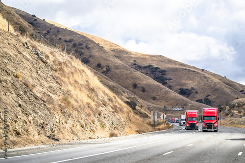 Team of two red big rig semi trucks transporting cargo in dry van semi trailers driving uphill on the mountain highway road in California