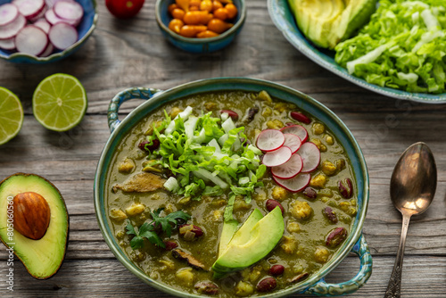 Vegan Pozole Verde dish with hominy, beans, tomatillo, and button mushrooms topped with avocado, radish, and shredded napa cabbage. A spoon and more toppings on the side.  photo