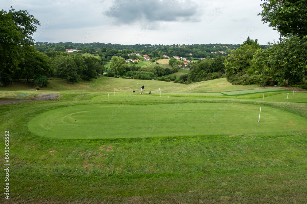 Campo de golfe rodeado por verdes campos a alguma floresta à volta com um golfista ao fundo a treinar as suas habilidades num dia muito nublado