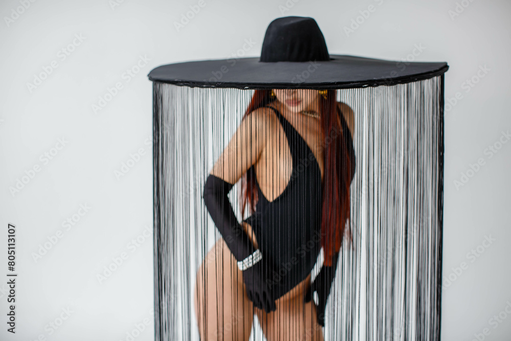 A portrait of a red-haired girl in a black bodysuit, gloves and a hat with threads stands in the studio on a white background.