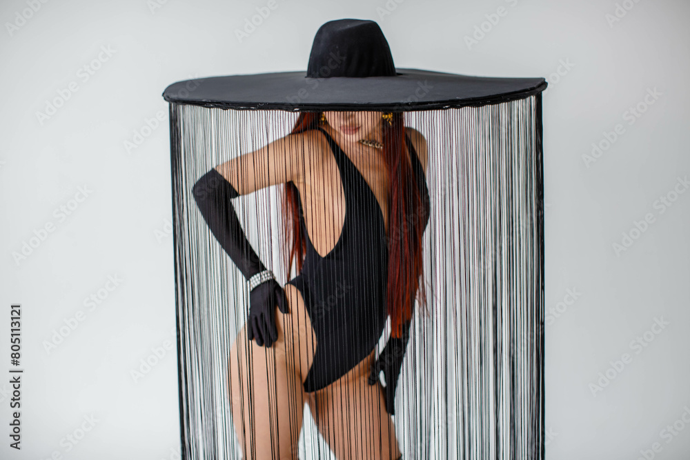 A portrait of a red-haired girl in a black bodysuit, gloves and a hat with threads stands in the studio on a white background.