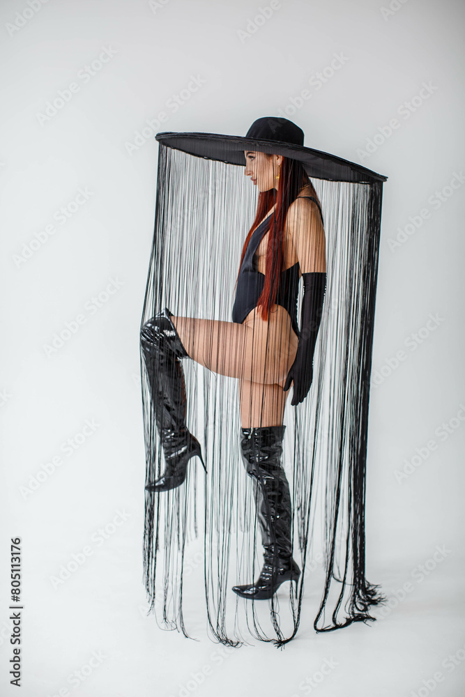 A red-haired girl in a black bodysuit, patent leather boots, gloves and a hat with threads stands in the studio on a white background