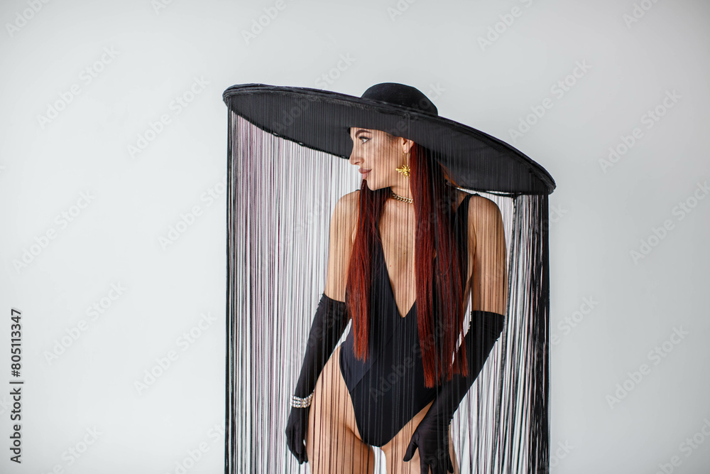 A portrait of a red-haired girl in a black bodysuit, gloves and a hat with threads stands in the studio on a white background.