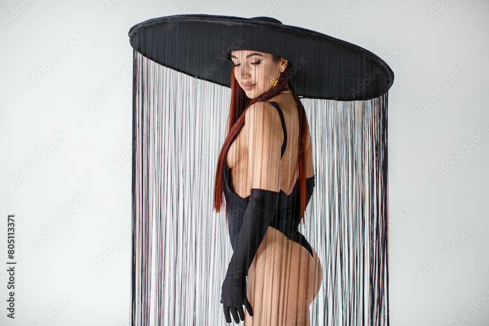 A portrait of a red-haired girl in a black bodysuit, gloves and a hat with threads stands in the studio on a white background.