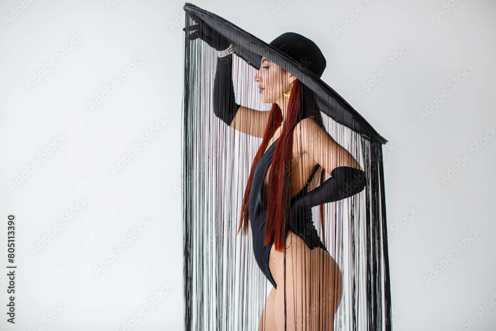 A portrait of a red-haired girl in a black bodysuit, gloves and a hat with threads stands in the studio on a white background.