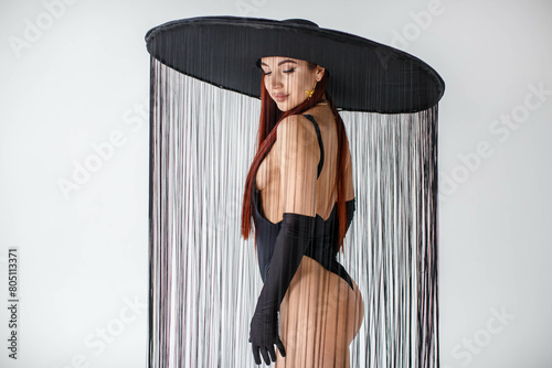 Obraz na płótnie A portrait of a red-haired girl in a black bodysuit, gloves and a hat with threads stands in the studio on a white background. w salonie