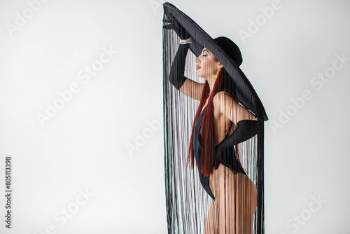 Obraz na płótnie A portrait of a red-haired girl in a black bodysuit, gloves and a hat with threads stands in the studio on a white background. w salonie