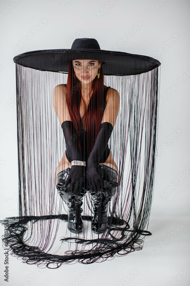 A red-haired girl in a black bodysuit, gloves, patent leather boots and a hat with threads sits in the studio on a white background.