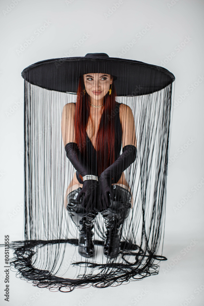 A red-haired girl in a black bodysuit, gloves, patent leather boots and a hat with threads sits in the studio on a white background.