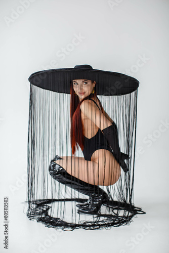 Obraz na płótnie A red-haired girl in a black bodysuit, gloves, patent leather boots and a hat with threads sits in the studio on a white background. w salonie