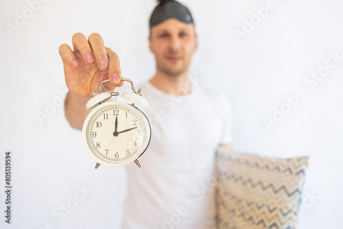 Young Caucasian man in pajamas holding alarm clock in his hand. Time management concept.