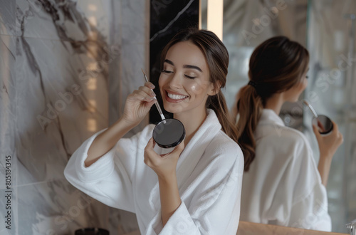 A beautiful woman in her bathroom  smiling and applying powder with the brush to one side of face  wearing white robe