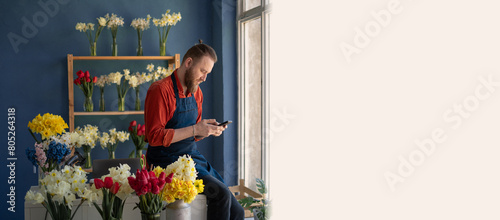 male self employed florist sitting at work desk focused on mobile phone screen. Man flower arranger reading email from customer photo