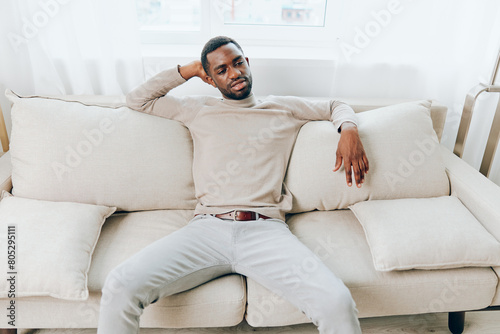Happy African American man sitting on a comfortable sofa in his modern living room, smiling and relaxing during the weekend He is alone, holding a smartphone and thinking, while enjoying the peace and