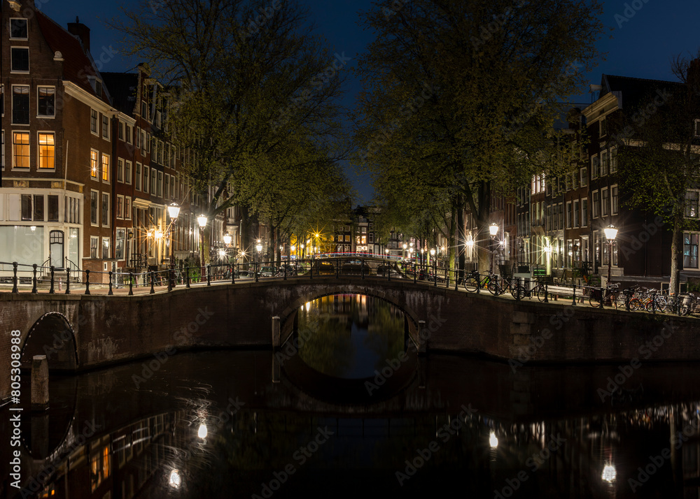 Naklejka premium Houses and bridges reflecting on the calm waters of the canals in Amsterdam at night with cars light trails