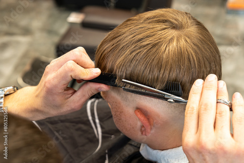 Man barber cutting little boy's hair using comb and scissors. Child getting haircut from adult male, likely barber. Professional hairdresser and cute client at modern barbershop. © BUCSA