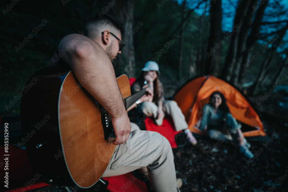A group of friends relax by a lakeside campsite, enjoying music from a guitar, sitting near an orange tent surrounded by trees.